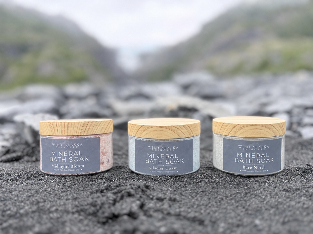 Three small jars of Wild Alaska Mineral Bath Soak with light wood lids are lined up on a dark textured surface with a blurred mountain landscape in the background. The labels show different scent names: Midnight Blossom, Glacier Coast, and Bare Roots.