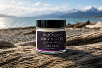 Jar of Wild Alaska Body Butter on a rocky beach with ocean and mountains in the background